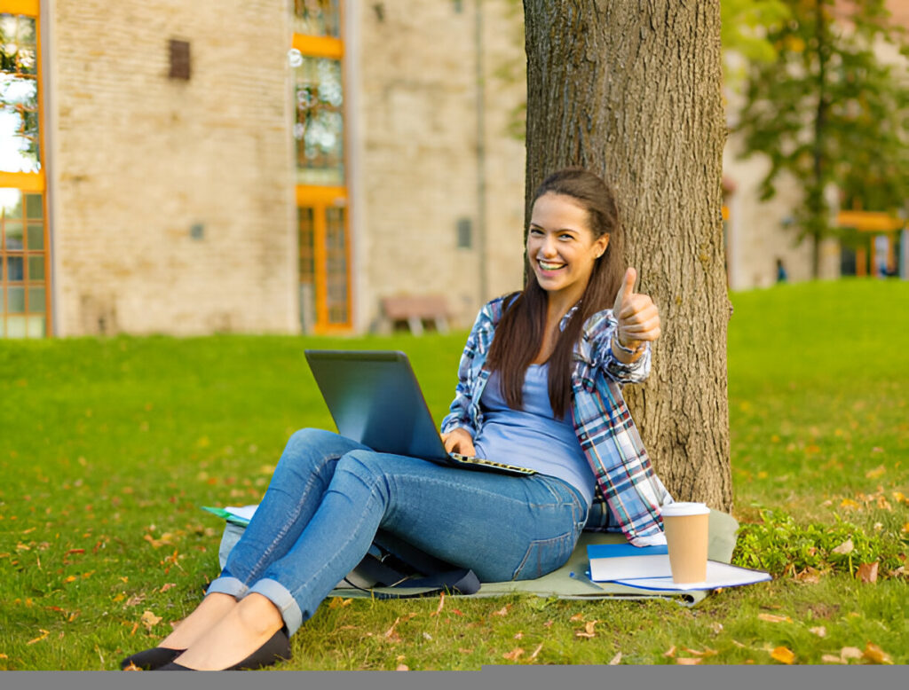 Happy student sitting under a tree with a laptop, giving a thumbs-up after receiving the Lumberjack Scholarship.