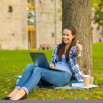 Happy student sitting under a tree with a laptop, giving a thumbs-up after receiving the Lumberjack Scholarship.