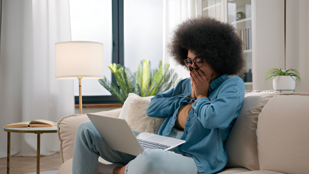 Young Black student with afro and glasses, looking surprised and joyful at her laptop, reacting to good news about her All Star Verified Scholarship application.